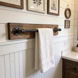 May include: A rustic towel rack made of dark stained wood and black metal pipes. A white towel hangs from the bar. The rack is mounted on a white paneled wall, creating a farmhouse style.