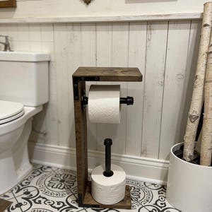 May include: A freestanding toilet paper holder constructed from dark stained wood and black industrial metal piping. The unit features a top shelf, a horizontal arm for a roll of toilet paper, and a vertical base post for storing an extra roll.
