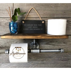 May include: A rustic wooden shelf with a metal pipe toilet paper holder. The shelf holds a roll of white toilet paper, a small vase with blue decorative stones and greenery, and a black sign that reads "MY HOUSE WAS TIDY LAST WEEK. SORRY YOU MISSED IT!"