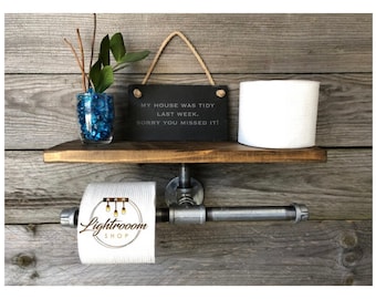 May include: A wall-mounted toilet paper holder made of industrial grey metal pipes with a dark brown wooden shelf. The shelf holds a white toilet paper roll, a glass vase with blue glass beads and green stems, and a black slate sign with the text MY HOUSE WAS TIDY LAST WEEK. SORRY YOU MISSED IT!