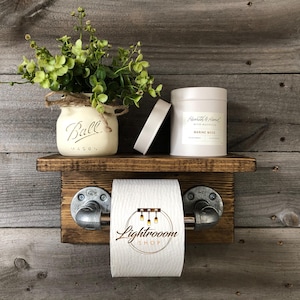 May include: Rustic wooden bathroom shelf with a toilet paper holder, a Ball Mason jar with greenery, a candle, and a small white object. The toilet paper holder has metal pipe fittings.