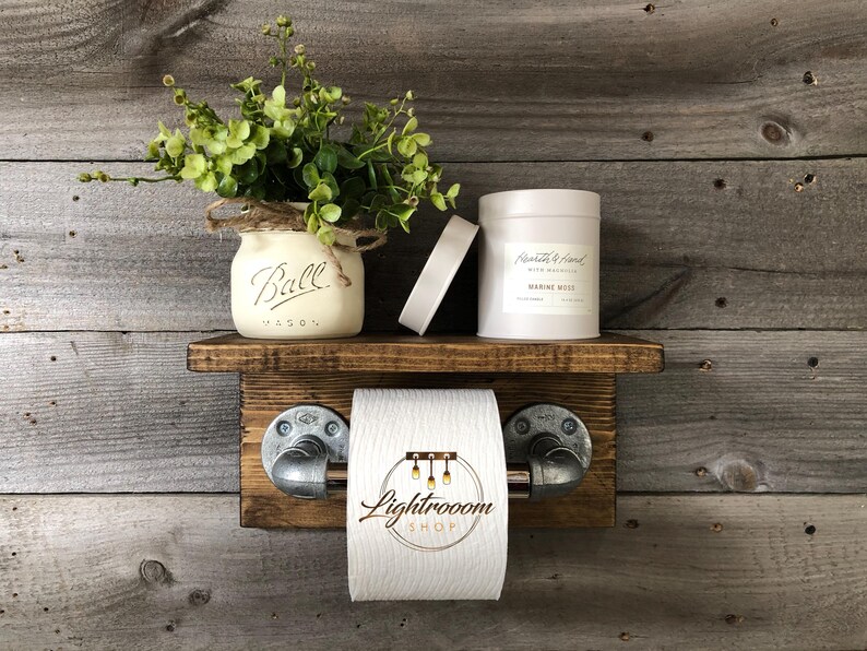 May include: Rustic wooden bathroom shelf with a toilet paper holder, a Ball Mason jar with greenery, a candle, and a small white object. The toilet paper holder has metal pipe fittings.