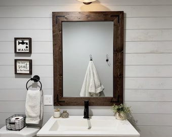 May include: A rectangular bathroom mirror with a dark brown wooden frame featuring black metal corner accents. The mirror is mounted on a white shiplap wall above a white vanity sink, a black faucet, a white towel, and two small wall signs.