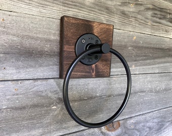 May include: A wall-mounted towel ring featuring a circular black metal hoop attached to a dark brown square wooden base. The metal hardware is secured with four visible screws, showcasing a matte black finish on the ring and mounting plate.