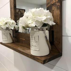 May include: A wooden shelf with a dark brown frame and a mirror. Two white ceramic vases with white flowers sit on the shelf. The vases have a metal rim and a rope handle. The text "HOME your story begins" is printed on the vases.