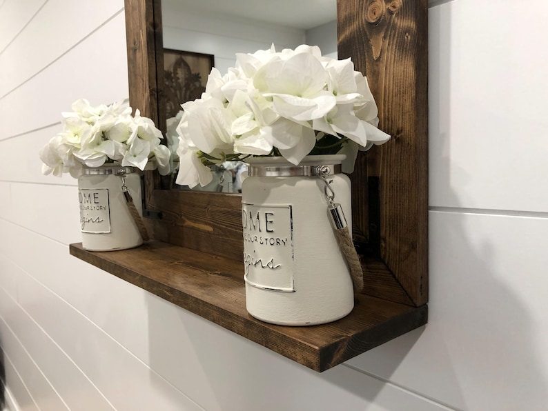 May include: A wooden shelf with a dark brown frame and a mirror. Two white ceramic vases with white flowers sit on the shelf. The vases have a metal rim and a rope handle. The text "HOME your story begins" is printed on the vases.