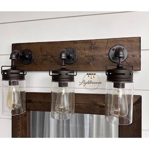 May include: A three-light vanity fixture mounted on a dark stained wooden board. It features black metal pipe hardware, cylindrical clear glass shades, and Edison-style bulbs. The text Lightrooom Shop is visible in the center of the fixture.