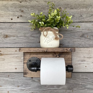 May include: Rustic wooden toilet paper holder with a shelf, featuring a black pipe design. A white mason jar with the word "Ball" is on the shelf, holding greenery. The holder is mounted on a weathered wood background.