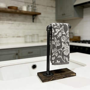 May include: A black metal pipe towel holder mounted on a rectangular dark brown wooden base. A grey hand towel with a white floral pattern hangs over the horizontal pipe bar. The structure features industrial-style fittings and a sturdy base.