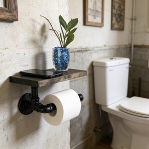 Puede incluir: Un soporte para papel higi&eacute;nico montado en la pared hecho de tuber&iacute;a industrial negra con un estante de madera oscura encima. El estante sostiene un tel&eacute;fono inteligente negro y un florero de vidrio transparente con piedras azules y una planta verde. Un rollo de papel blanco est&aacute; colocado.