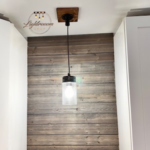 May include: A pendant light fixture with a clear glass shade and a black metal cap. The light hangs from a black cord attached to a wooden square. The fixture is mounted on a white ceiling and a wooden plank wall.