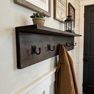 May include: A dark brown wooden wall-mounted coat rack featuring a flat top shelf and five black metal hooks. The rack is constructed from horizontal planks and is secured to the wall with black metal brackets. A tan jacket hangs from one hook.