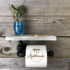 Puede incluir: Un soporte de papel higi&eacute;nico montado en la pared con un estante de madera gris sostenido por tuber&iacute;as de metal industrial negro. Un rollo de papel higi&eacute;nico blanco con el texto Lightrooom SHOP impreso cuelga de la tuber&iacute;a. Un jarr&oacute;n de vidrio con piedras azules y una planta descansa sobre el estante.