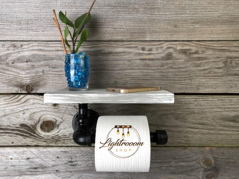 Puede incluir: Un soporte de papel higi&eacute;nico montado en la pared con un estante de madera gris sostenido por tuber&iacute;as de metal industrial negro. Un rollo de papel higi&eacute;nico blanco con el texto Lightrooom SHOP impreso cuelga de la tuber&iacute;a. Un jarr&oacute;n de vidrio con piedras azules y una planta descansa sobre el estante.