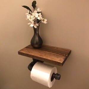 Puede incluir: Un soporte de papel higi&eacute;nico montado en la pared con un estante de madera marr&oacute;n oscuro sostenido por tuber&iacute;as de metal industrial negro. Un rollo de papel higi&eacute;nico blanco est&aacute; montado en la tuber&iacute;a y un jarr&oacute;n oscuro con flores blancas descansa sobre el estante.