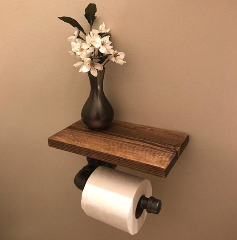 Puede incluir: Un soporte de papel higi&eacute;nico montado en la pared con un estante de madera marr&oacute;n oscuro sostenido por tuber&iacute;as de metal industrial negro. Un rollo de papel higi&eacute;nico blanco est&aacute; montado en la tuber&iacute;a y un jarr&oacute;n oscuro con flores blancas descansa sobre el estante.