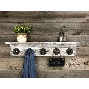 May include: A rustic wooden shelf with a distressed white finish, featuring five black metal hooks. A blue scarf hangs from one hook. The shelf holds a mason jar with greenery, a glass spray bottle, and a small sign that reads "MY HOUSE WAS TIDY LAST WEEK. SORRY YOU MISSED IT!"