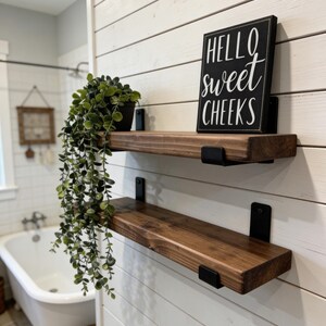 May include: Two wooden shelves with black metal brackets are mounted on a white shiplap wall. A small potted plant sits on the top shelf, and a black sign with the words "Hello Sweet Cheeks" is on the top shelf.