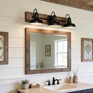 May include: A three-light vanity fixture with matte black metal dome shades mounted on a horizontal dark wood plank, positioned above a rectangular mirror with a matching dark wood frame, featuring a white sink and black faucet hardware.