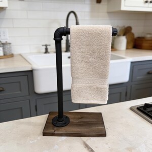 May include: A freestanding towel holder constructed from black industrial metal piping attached to a rectangular dark brown wooden base. A single beige hand towel is draped over the horizontal pipe arm, which features a threaded end cap.