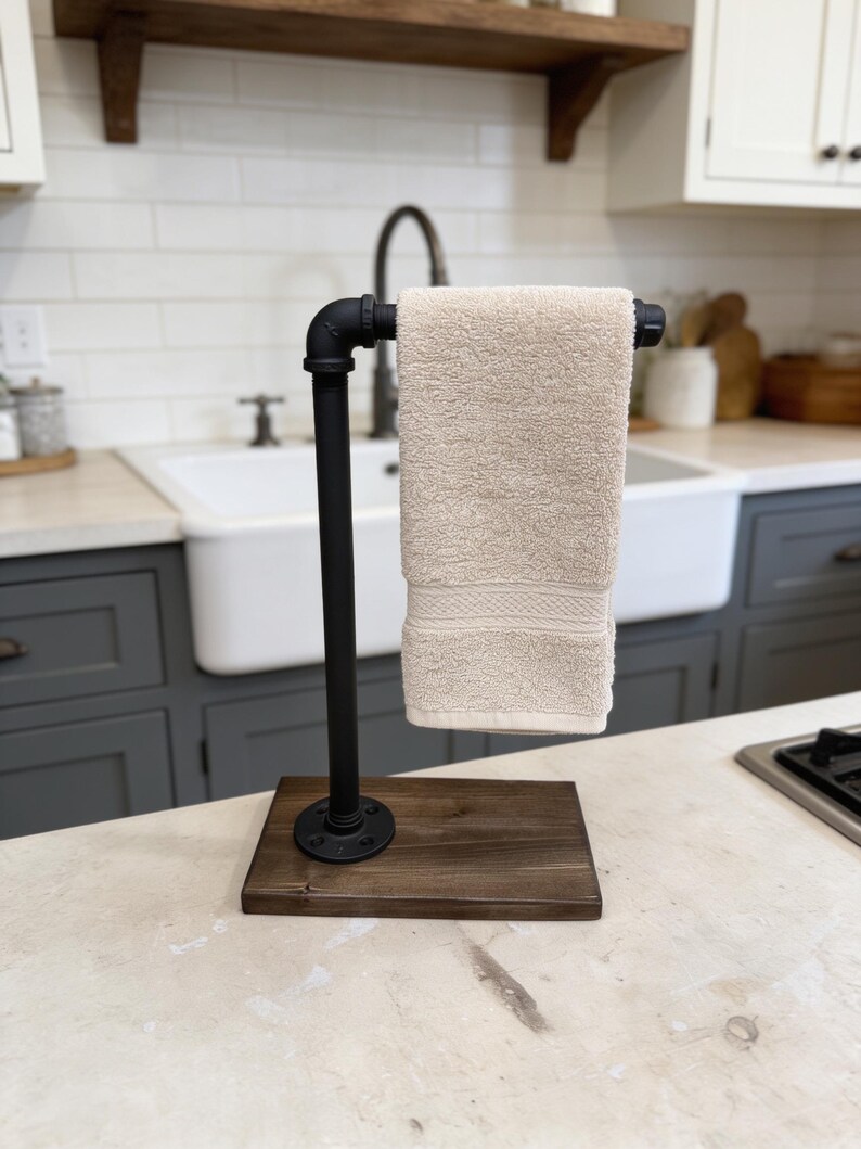 May include: A freestanding towel holder constructed from black industrial metal piping attached to a rectangular dark brown wooden base. A single beige hand towel is draped over the horizontal pipe arm, which features a threaded end cap.