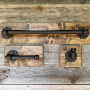 Puede incluir: Un juego de tres accesorios de ba&ntilde;o con tuber&iacute;as de metal negro mate montadas sobre tablas de madera te&ntilde;ida rectangulares. El conjunto incluye una barra larga para toallas, un portarrollos de papel higi&eacute;nico y un gancho de pared.