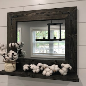 May include: A rustic, square wooden mirror with a dark stain and metal corner brackets. A shelf below the mirror holds cotton stems in a mason jar and loose cotton bolls. The mirror reflects a window and light fixture.