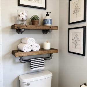 May include: Two wall-mounted wooden shelves with dark metal pipe brackets. The top shelf holds a glass bowl with cotton balls, a small succulent in a terracotta pot, and a blue and white soap dispenser. The bottom shelf holds three white towels and a candle.