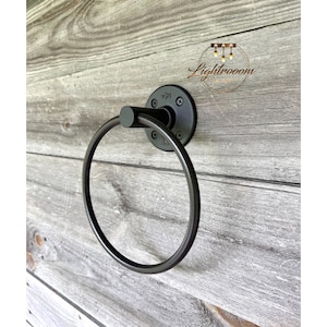 May include: A black metal towel ring mounted on a wooden wall. The ring is circular and attached to a round base with visible screws. The background is made of weathered wooden planks.