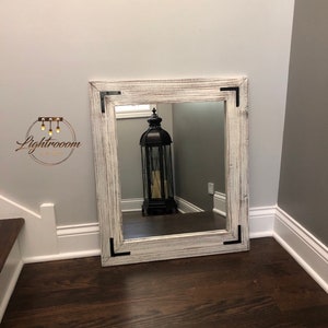 May include: A large, square mirror with a distressed white wooden frame and black corner accents. The mirror is leaning against a gray wall on a dark wood floor. A black lantern is reflected in the mirror.