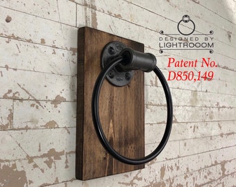 May include: A wall-mounted towel ring featuring a dark brown rectangular wooden base and a black metal pipe fitting. The ring is attached to a circular flange secured to the wood. Text on the wall reads DESIGNED BY LIGHTROOOM and Patent No. D850,149.