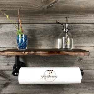 May include: A rustic, dark-stained wooden shelf with black pipe brackets, holding a white paper towel roll. A glass spray bottle and a small vase with blue decorative stones and greenery are on the shelf. The shelf is mounted on a wooden wall.
