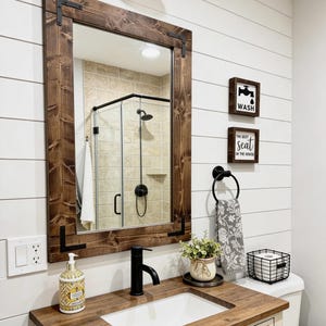May include: A bathroom vanity with a dark wood countertop, white sink, and black faucet. Above is a rectangular mirror with a dark wood frame and black metal corner brackets. Two square wall signs read WASH and THE BEST seat IN THE HOUSE.