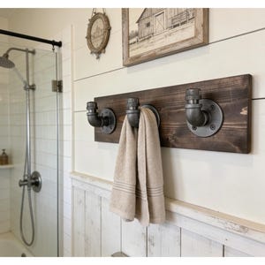 May include: A wall-mounted towel rack featuring a dark brown rectangular wooden board with three industrial-style gray metal pipe hooks. A beige towel hangs from the center hook. The hardware is secured with circular metal flanges and screws.
