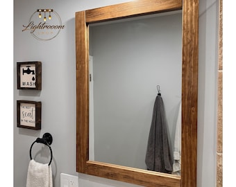May include: A rectangular mirror with a thick, brown wooden frame hangs on a gray wall. Below the mirror are decorative items, including a towel ring with a white towel, a soap dispenser, and two small framed signs. The top sign says "Wash".