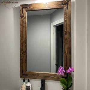 May include: A rectangular wall mirror featuring a dark brown stained wood frame with visible grain and black metal corner brackets. The mirror is mounted above a white sink basin with a black faucet, two soap dispensers, and a potted orchid.