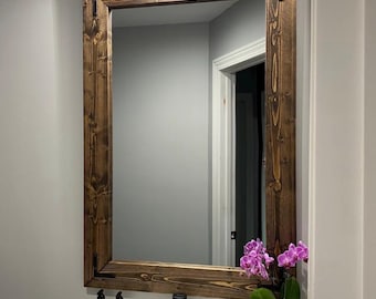 May include: A rectangular wall mirror featuring a dark brown stained wood frame with visible grain and black metal corner brackets. The mirror is mounted above a white sink basin with a black faucet, two soap dispensers, and a potted orchid.