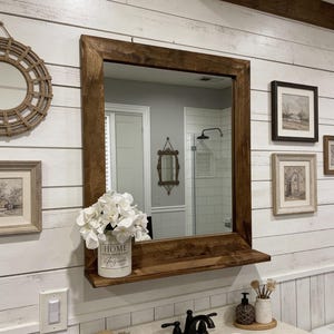 May include: A bathroom vanity with a large, square, dark wood-framed mirror. A shelf below the mirror holds a white vase with white flowers and the word "HOME" printed on it. The vanity has a white countertop and a black faucet.