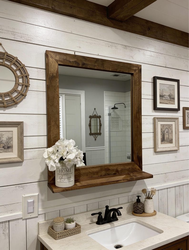 May include: A bathroom vanity with a large, square, dark wood-framed mirror. A shelf below the mirror holds a white vase with white flowers and the word "HOME" printed on it. The vanity has a white countertop and a black faucet.