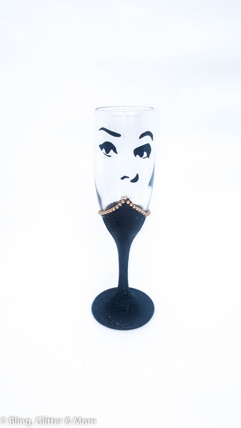 K&ouml;nnte beinhalten: Eine schwarze Champagnerfl&ouml;te mit einem goldenen Glitzerrand und einem schwarz-wei&szlig;en Gesichtsdesign. Das Gesicht hat ein zwinkerndes Auge und ein verschmitztes L&auml;cheln.