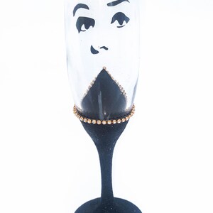 K&ouml;nnte beinhalten: Eine schwarze Champagnerfl&ouml;te mit goldfarbenem Glitzer am Rand und einem schwarz-wei&szlig;en Gesichtsdesign. Das Gesicht hat einen &uuml;berraschten Ausdruck mit gro&szlig;en Augen und einem kleinen Mund.