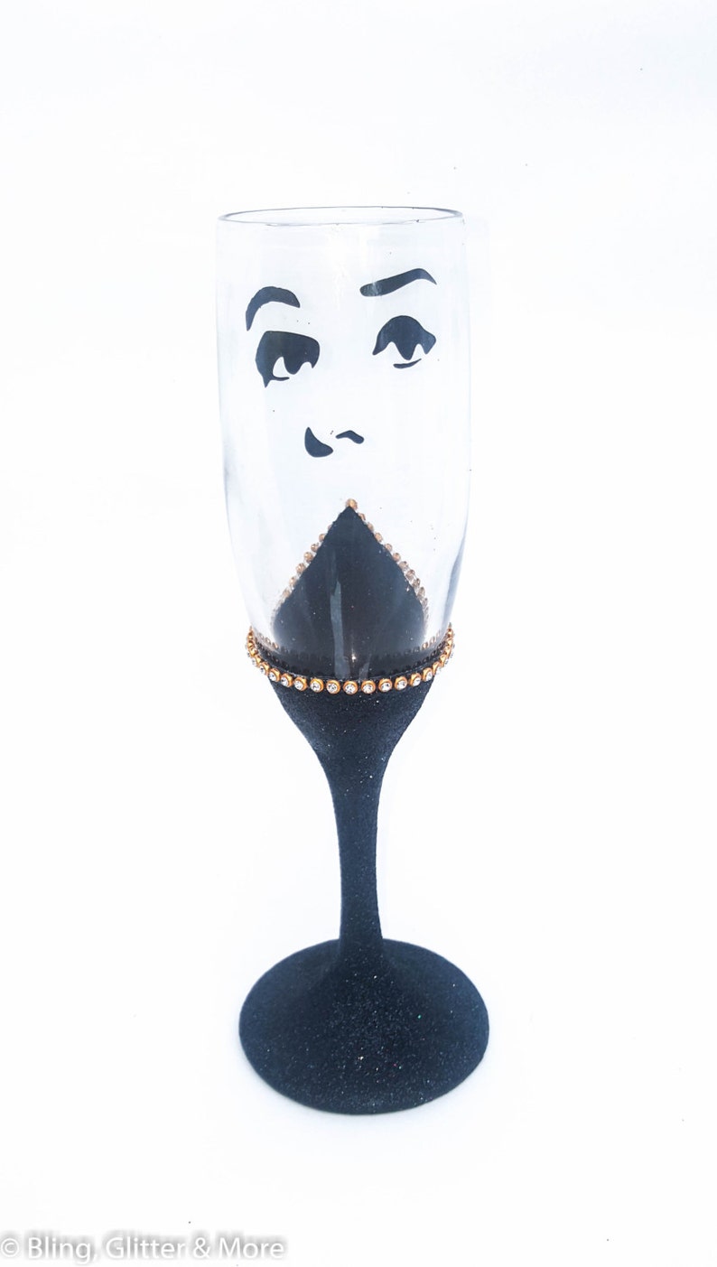 K&ouml;nnte beinhalten: Eine schwarze Champagnerfl&ouml;te mit goldfarbenem Glitzer am Rand und einem schwarz-wei&szlig;en Gesichtsdesign. Das Gesicht hat einen &uuml;berraschten Ausdruck mit gro&szlig;en Augen und einem kleinen Mund.