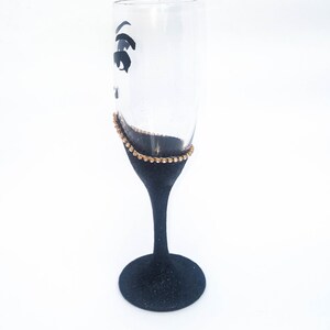 K&ouml;nnte beinhalten: Eine schwarze Champagnerfl&ouml;te mit einem goldenen Glitzerrand und einem schwarzen Augenmotiv. Die Fl&ouml;te steht auf einem wei&szlig;en Hintergrund.