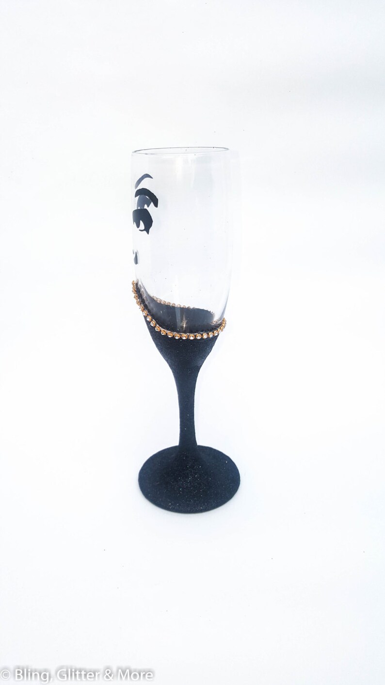 K&ouml;nnte beinhalten: Eine schwarze Champagnerfl&ouml;te mit einem goldenen Glitzerrand und einem schwarzen Augenmotiv. Die Fl&ouml;te steht auf einem wei&szlig;en Hintergrund.