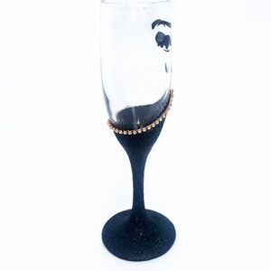 K&ouml;nnte beinhalten: Eine schwarze Champagnerfl&ouml;te mit goldenen Glitzer am Rand. Die Fl&ouml;te ist mit einer schwarzen Silhouette eines Frauenauges verziert.