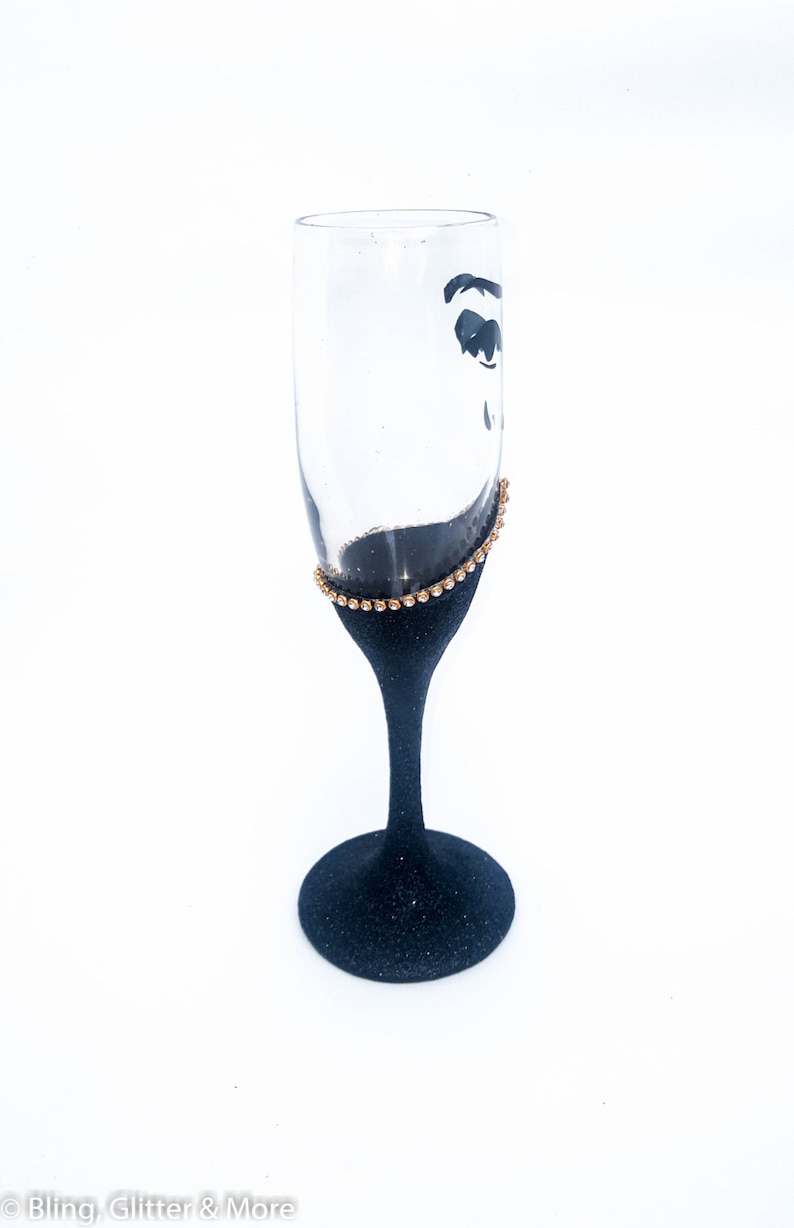 K&ouml;nnte beinhalten: Eine schwarze Champagnerfl&ouml;te mit goldenen Glitzer am Rand. Die Fl&ouml;te ist mit einer schwarzen Silhouette eines Frauenauges verziert.