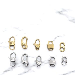 May include: A collection of gold and silver-toned lobster claw clasps and spring clasps. The clasps are in various sizes and shapes, perfect for jewelry making or crafting projects. The clasps are displayed on a white marble surface.