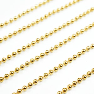 May include: A close-up of a gold ball chain. The chain is made of small, round beads that are evenly spaced. The chain is lying flat on a white surface.