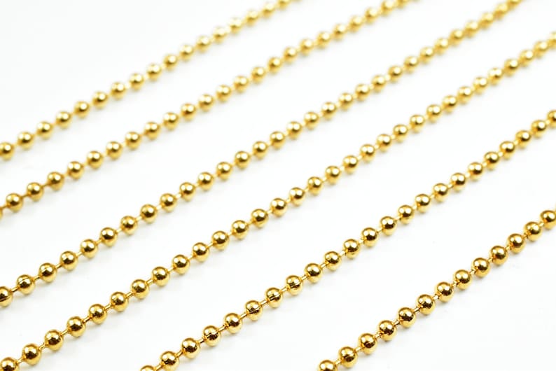 May include: A close-up of a gold ball chain. The chain is made of small, round beads that are evenly spaced. The chain is lying flat on a white surface.
