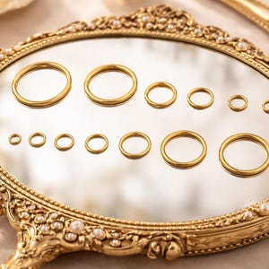 May include: A collection of twelve gold-toned metal rings of varying diameters arranged in two rows on a reflective surface. The rings are displayed on an ornate, oval-shaped gold frame decorated with small white pearls along its edge.